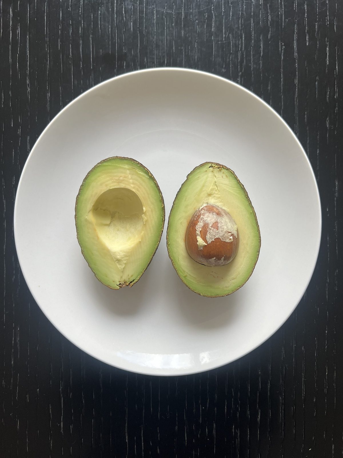 A ripe avocado cut in half on a white plate, one half holding the pit, with smooth pale-green flesh