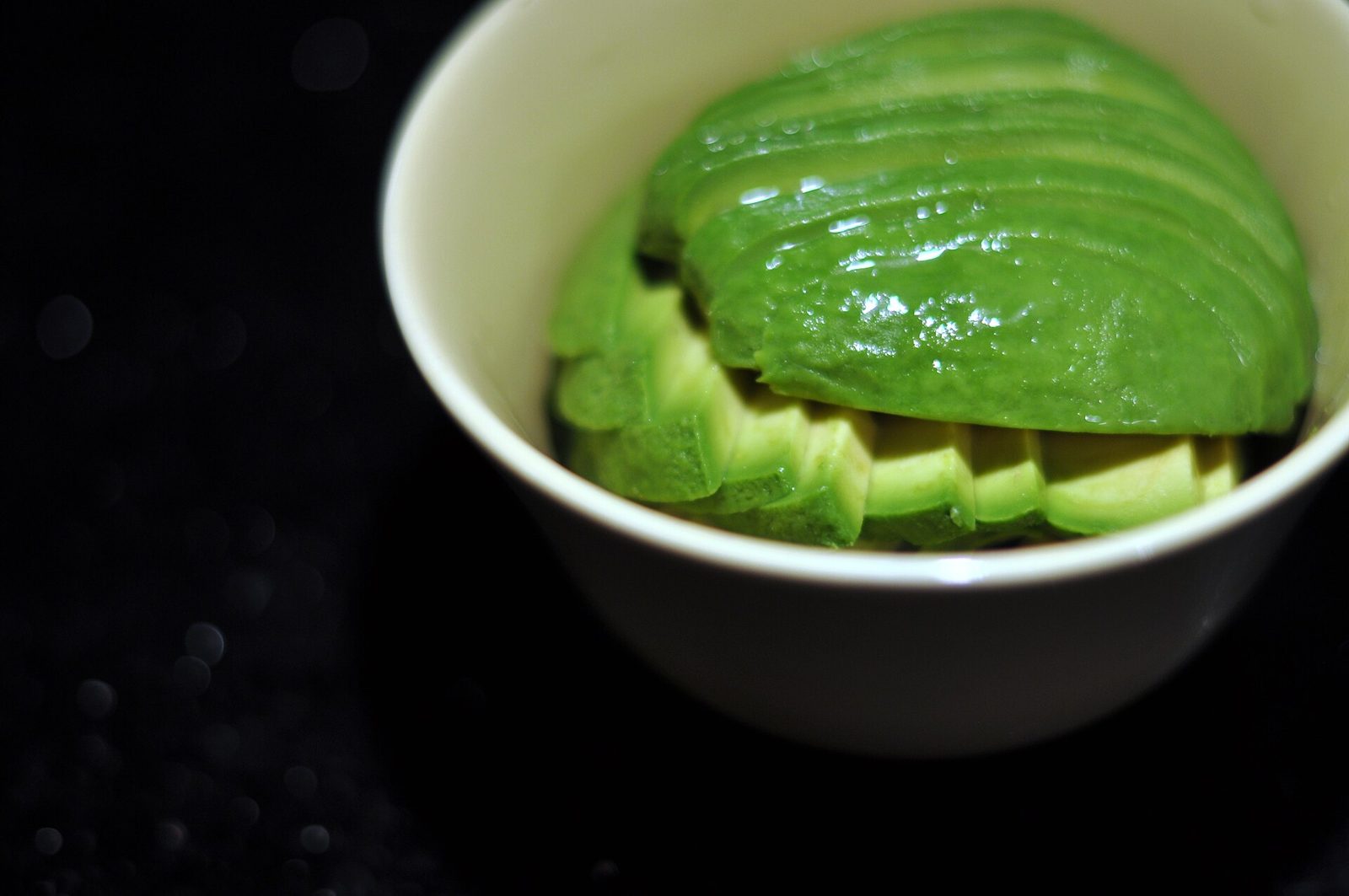 Thin slices of green avocado fanned in a small white bowl against a dark background