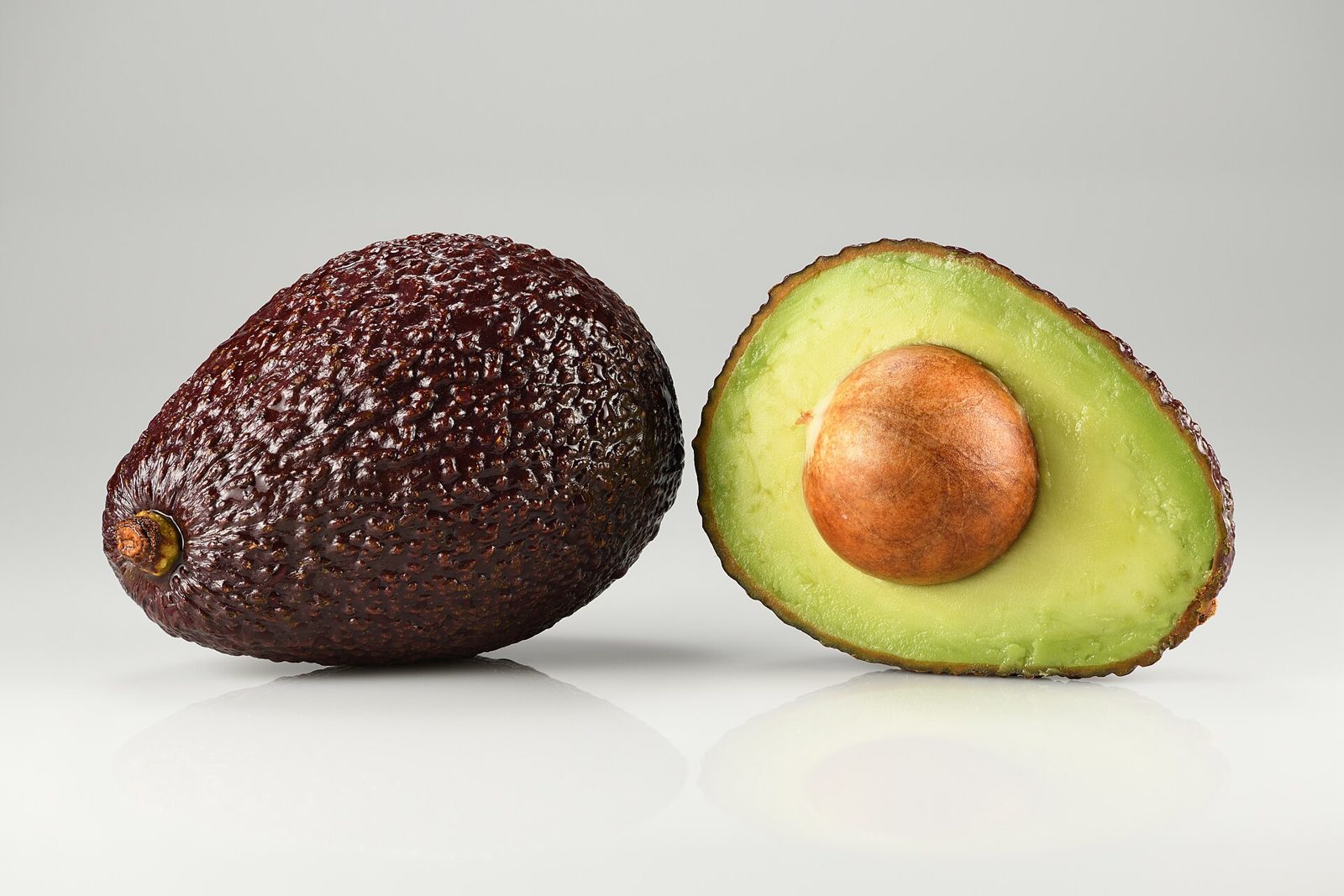 A whole dark pebbled Hass avocado beside a halved one showing green flesh and a round brown pit
