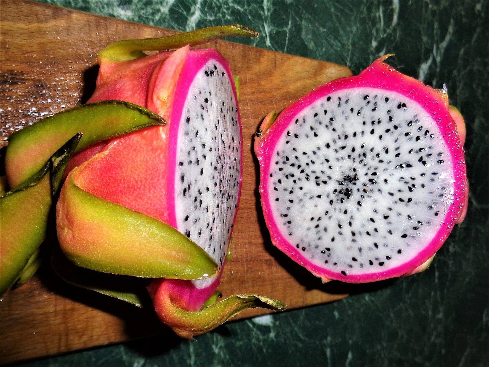 Two halves of a white-fleshed dragon fruit on a wooden board, one cut lengthwise and one across, showing white flesh with black seeds inside a bright pink rim