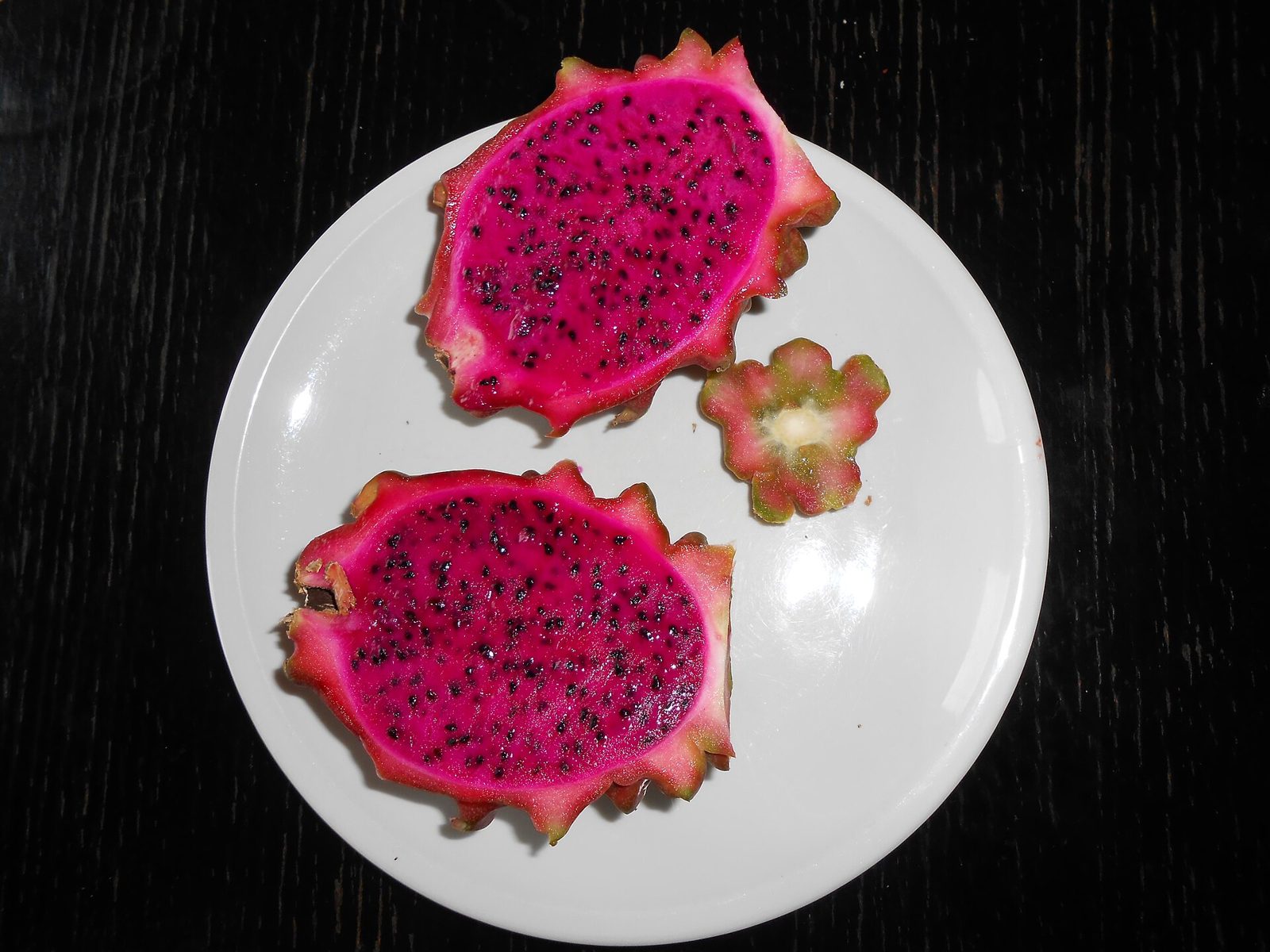 A red-fleshed dragon fruit cut in half on a white plate, showing magenta flesh with small black seeds, with a slice of the stem end beside it