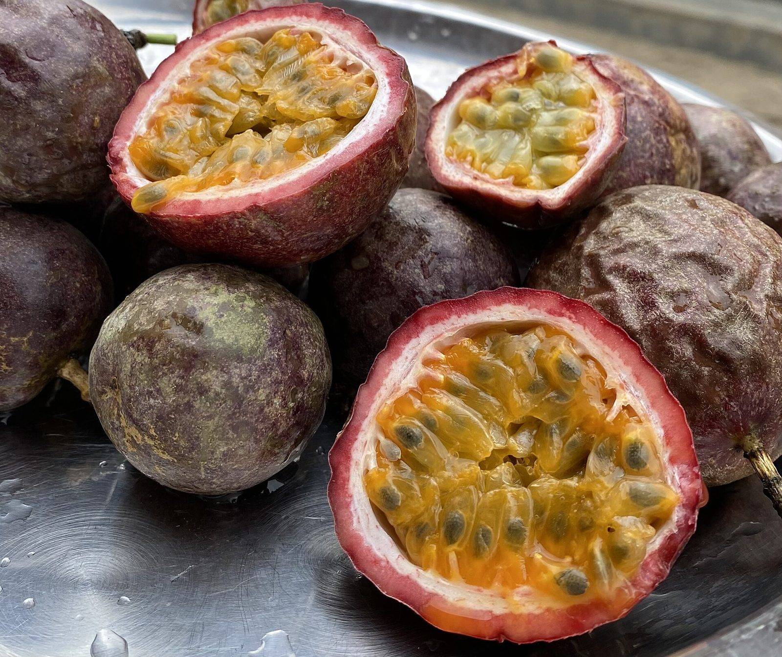 Several purple passion fruits on a metal tray, some whole with dull skin and some halved to show a pink rind, white pith and orange pulp with dark seeds