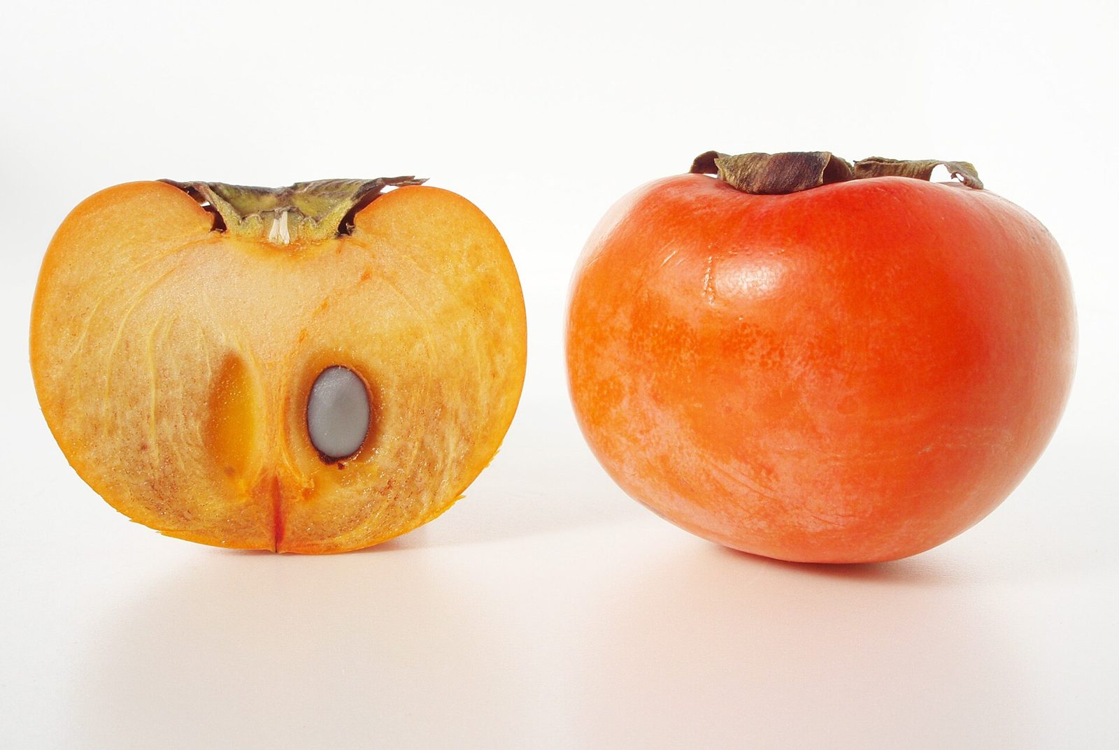 A Fuyu persimmon cut in half, showing orange flesh and a single dark seed, beside a whole orange-red persimmon
