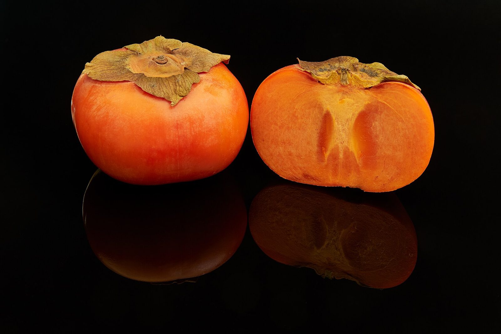 Two deep orange Fuyu persimmons on a black reflective surface, one whole with its dry calyx and one cut open to show the flesh