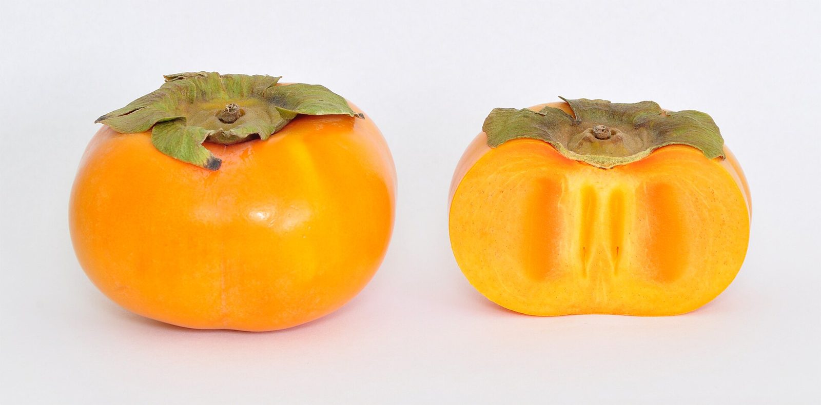 A whole orange persimmon with a green calyx beside another persimmon cut in half across the middle, showing orange flesh