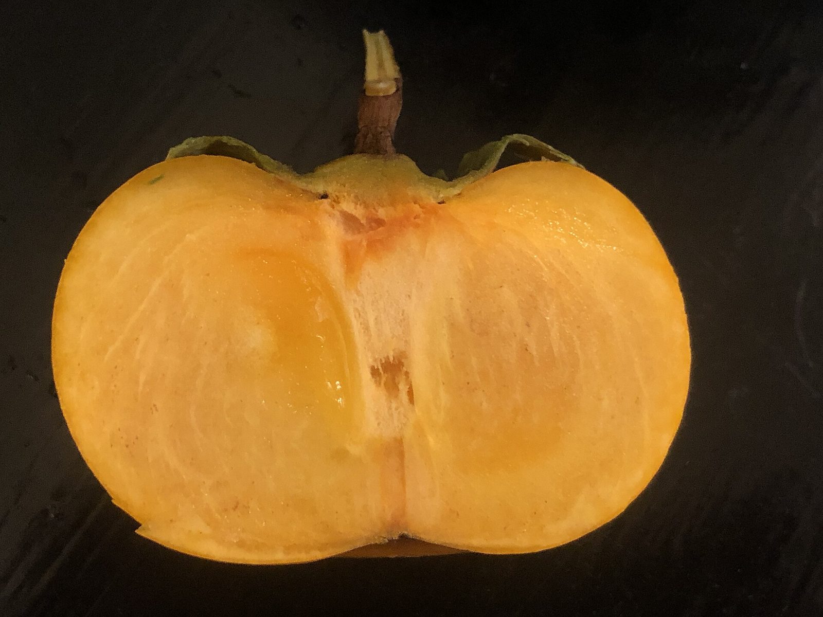 An Asian persimmon cut in half across the middle, showing smooth orange flesh with the stem and calyx on top