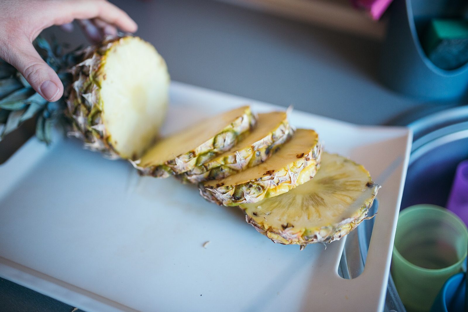 A pineapple with its crown still attached, cut into thick rounds on a white cutting board, with a hand steadying the uncut end