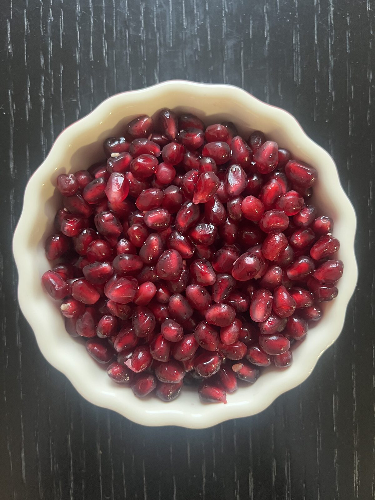 A shallow white bowl filled with glossy deep red pomegranate arils, seen from above on a dark wooden table